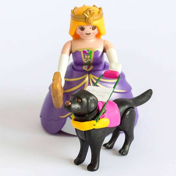 #ToyLikeMe makeover image showing Playmobil princess figure with a guide dog. The guide dog has a pink harness.