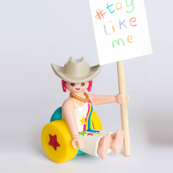 #ToyLikeMe makeover image showing Playmobil cowgirl figure using a model wheelchair. She is holding a sign which reads 