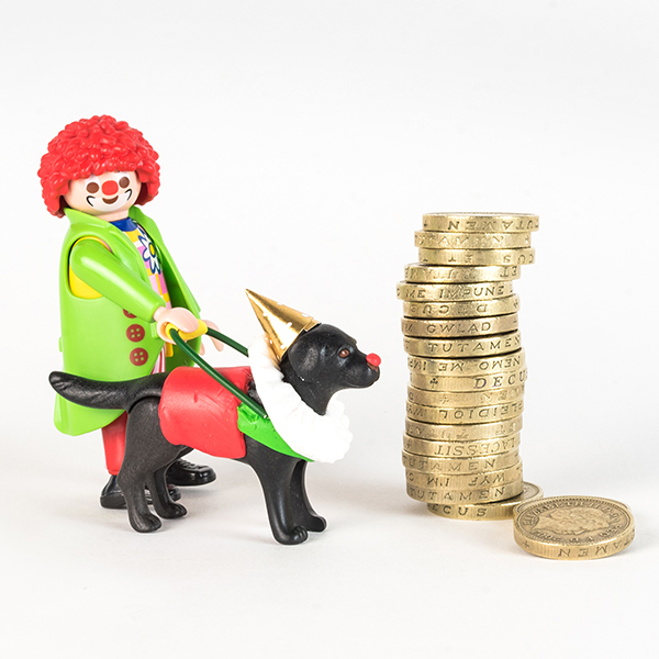 #ToyLikeMe makeover showing Playmobil clown figure with black lab guide dog also dressed in clown clothes. They are stood next to a stack of pound coins.