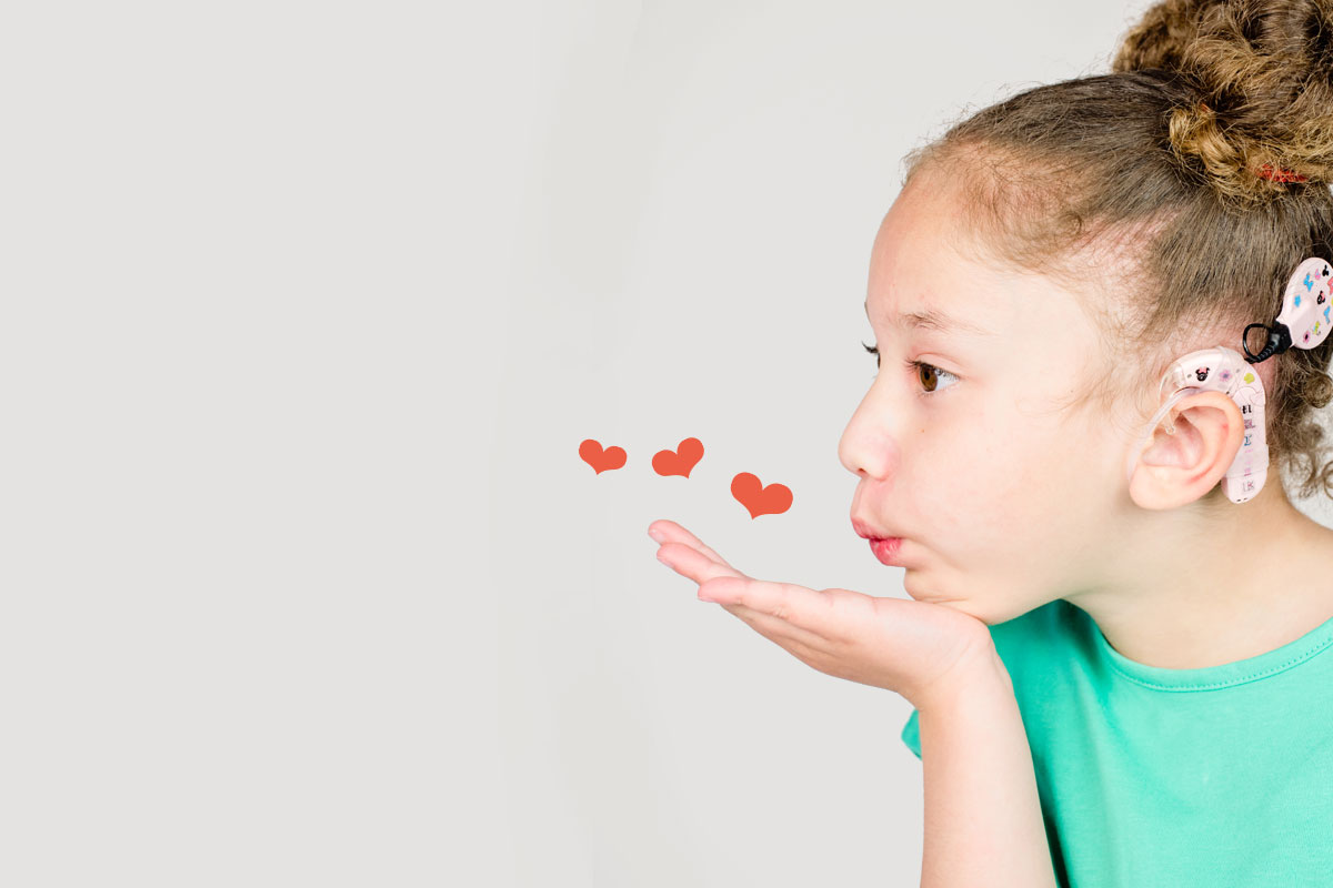 Image shows girl with cochlear implant blowing a kiss. There are hearts around her hand.