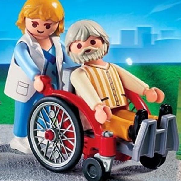 Image shows Playmobil wheelchair set featuring a red wheelchair. The man using the chair is an older man with grey hair and beard. There is a younger woman pushing the chair behind him.