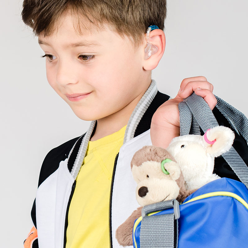 Image shows boy holding two plush toys. The boy and the toys all have hearing aids.