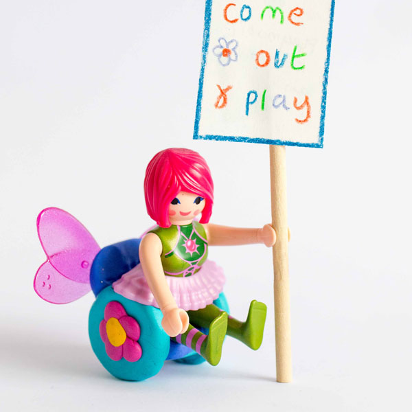 Image shows a #ToyLikeMe makeover of a Playmobil fairy figure. The fairy is sitting in a wheelchair made of modelling clay with a flower on the wheel and wings on the back. The fairy is holding a placard which reads 