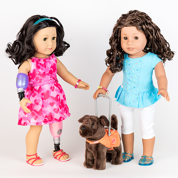 Two American Girl dolls stood next to one another. One has a prosthetic hand and arm, the other has a guide dog.