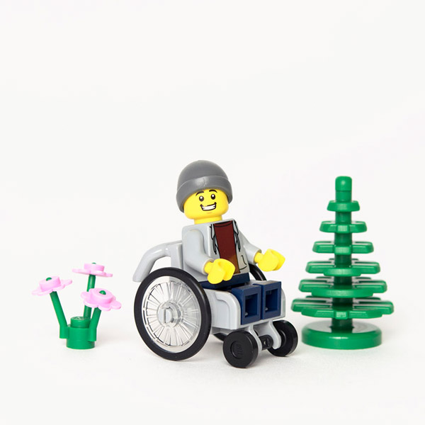 Lego mini-figure in grey hoodie and beaning hat using a grey wheelchair, sat next to Lego tree and flowers.