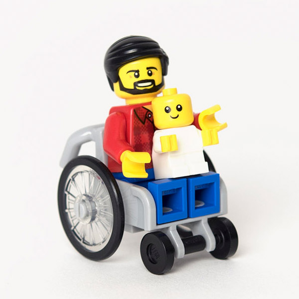 Lego dad mini-figure with red shirt and beard using a grey Lego wheelchair. Mini-figure baby is sat on his knew.