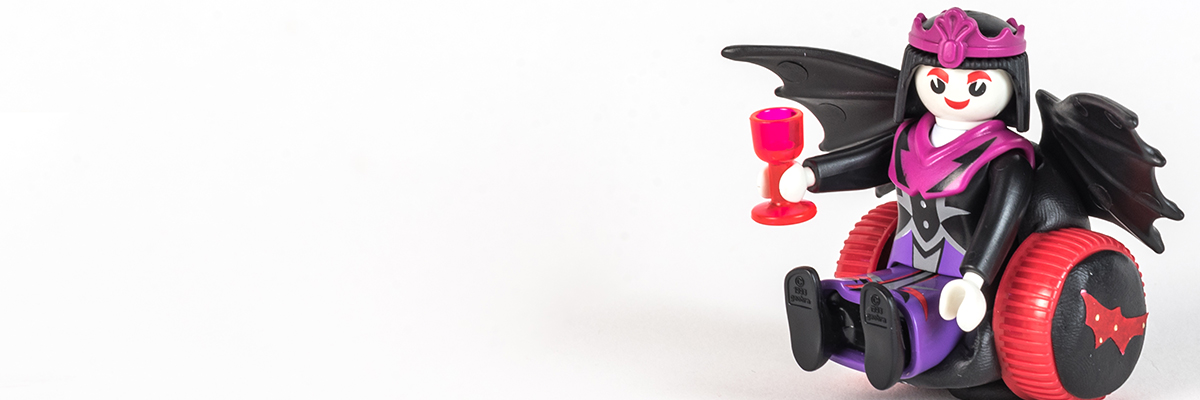 #ToyLikeMe makeover image of Playmobil Draculette figure using a red and black wheelchair made of modelling clay. The back of the chair has bat wings.