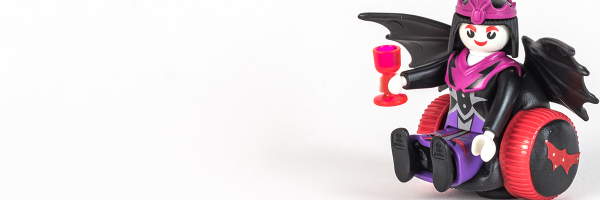 #ToyLikeMe makeover image of Playmobil Draculette figure using a red and black wheelchair made of modelling clay. The back of the chair has bat wings.