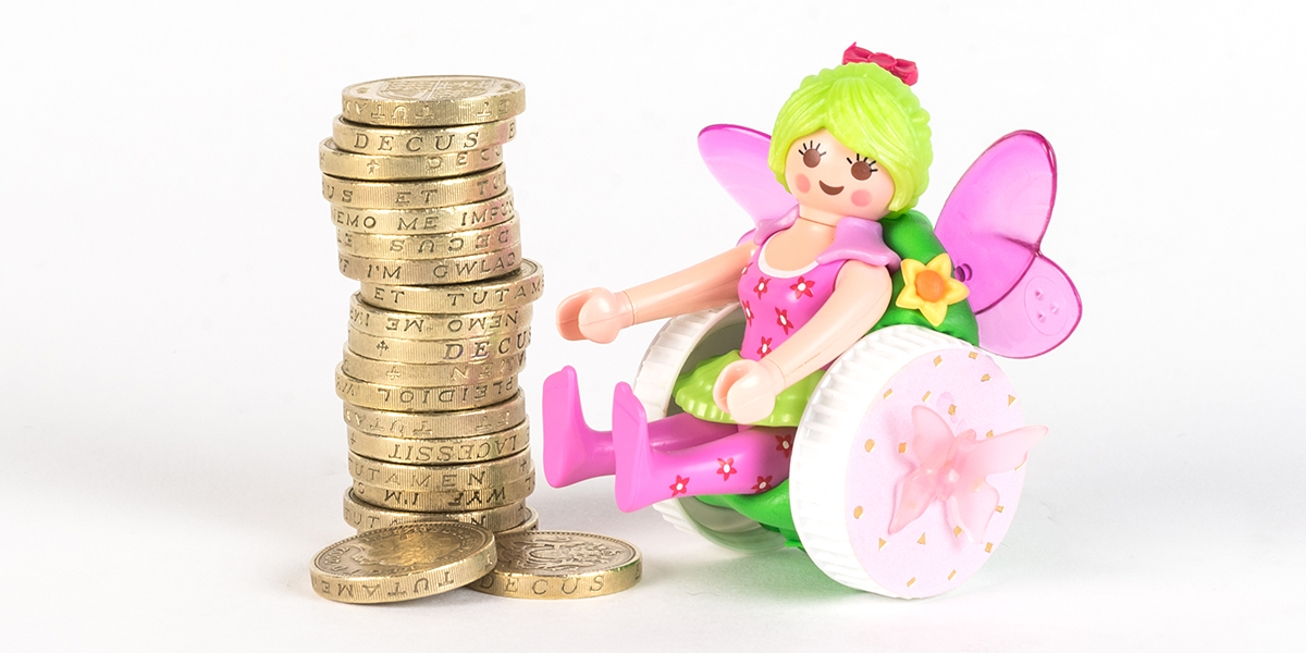 #ToyLikeMe makeover image of Playmobil fairy figure using a pink and green wheelchair made of modelling clay. The back of the chair has pink wings. She is sat next to a pile of one pound coins.
