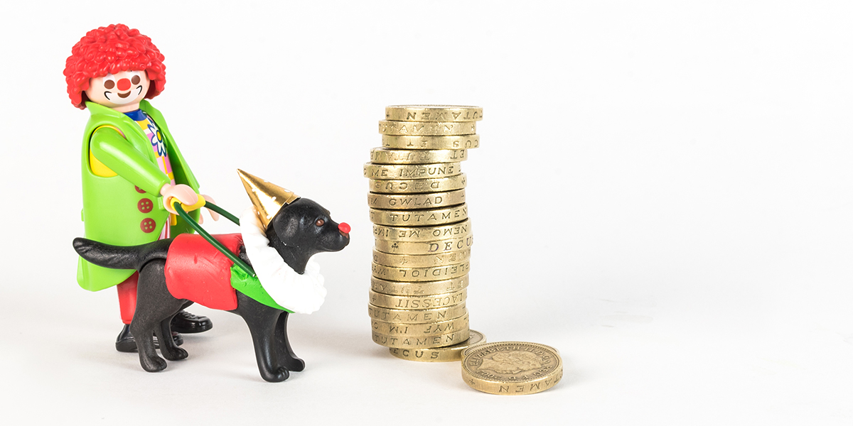 #ToyLikeMe makeover image of Playmobil clown figure with black guide dog. The dog is wearing a model clown's ruffle, gold pointy hat, red guide dog harness and has a red nose. They are stood next to a stack of one pound coins.