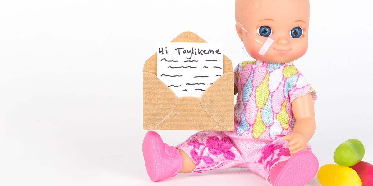 #ToyLikeMe makeover image showing baby girl toy with nasal feeding tube. The doll holding a little envelope.