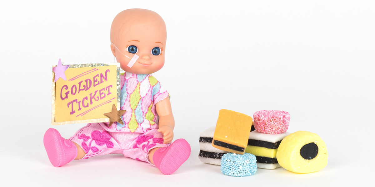 #ToyLikeMe makeover image showing baby girl toy with nasal feeding tube. The doll is sitting next to a pile of All Sorts sweets and holding a golden ticket.