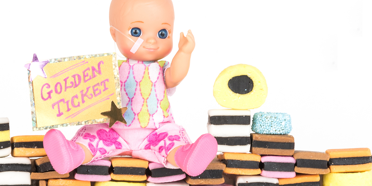 #ToyLikeMe makeover image showing baby girl toy with nasal feeding tube. The doll is sitting on a wall made of All Sorts sweets and holding a golden ticket.