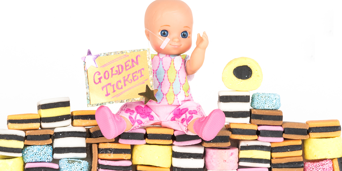 #ToyLikeMe makeover image showing baby girl toy with nasal feeding tube. The doll is sitting on a wall made of All Sorts sweets and holding a golden ticket.