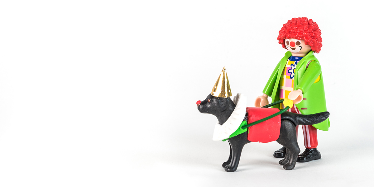 #ToyLikeMe makeover image of Playmobil clown figure with black guide dog. The dog is wearing a model clown's ruffle, gold pointy hat, red guide dog harness and has a red nose.