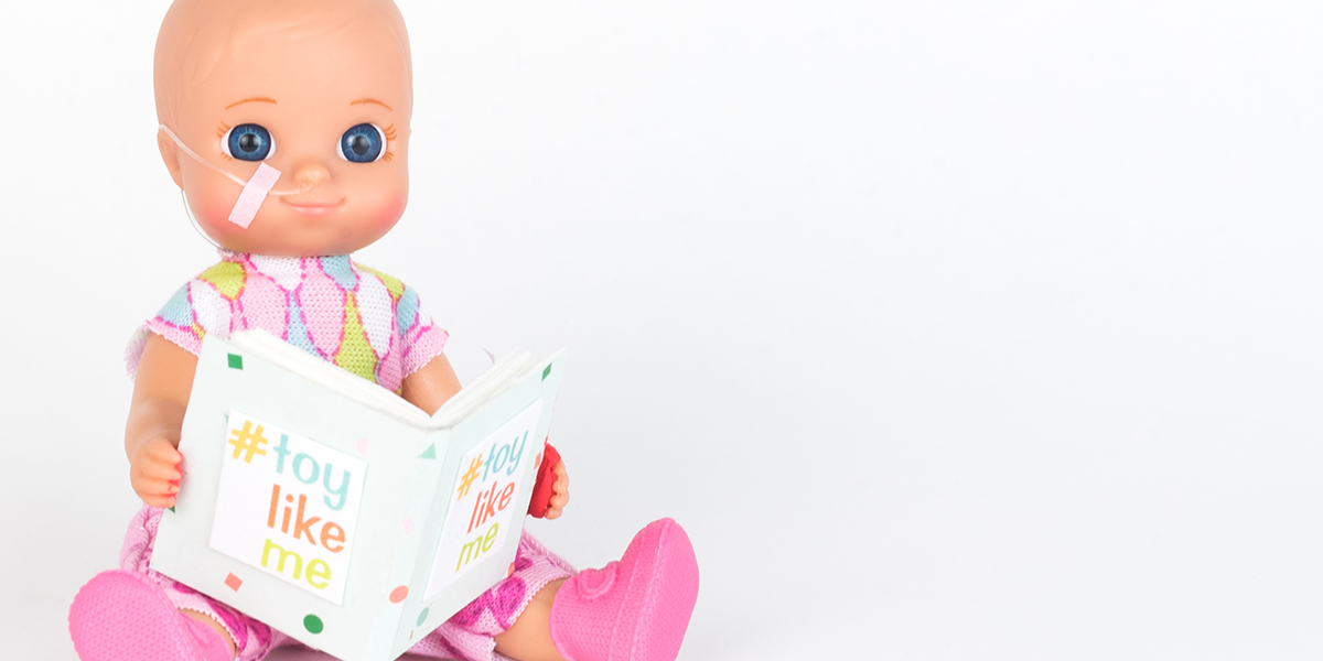 #ToyLikeMe makeover image showing baby girl toy with nasal feeding tube, reading a book.