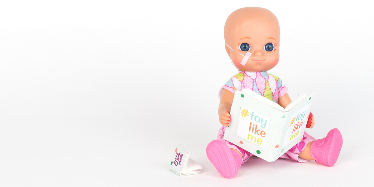 #ToyLikeMe makeover image showing baby girl toy with nasal feeding tube, reading a book.