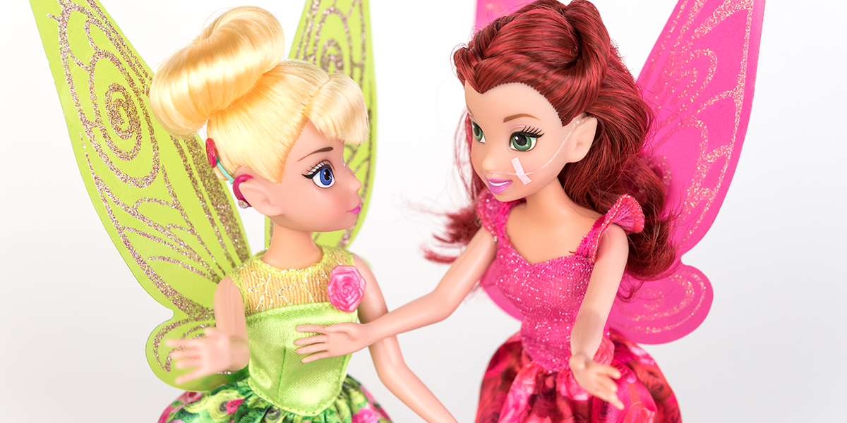 #ToyLikeMe makeover image showing Disney Tinkerbell doll with a hot pink model cochlear implant chatting with Disney fairy Rosetta who has a nasal feeding tube.