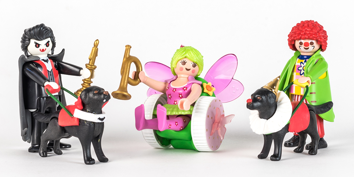 #ToyLikeMe makeover image showing a group of Playmobil figures. Dracula and a clown with guide dogs and a fairy with a wheelchair made of modelling clay.
