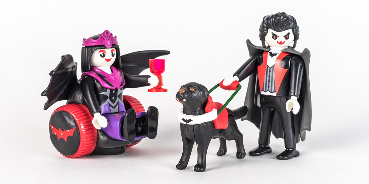 #ToyLikeMe makeover image of Playmobil Draculette figure using a red and black wheelchair made of modelling clay. The back of the chair has bat wings. Stood next to her is Dracula with a guide dog. The guide dog has fangs and a red harness with a bat on it.