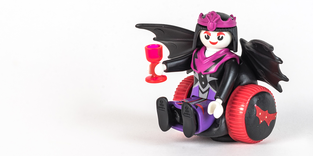 #ToyLikeMe makeover image of Playmobil Draculette figure using a red and black wheelchair made of modelling clay. The back of the chair has bat wings.