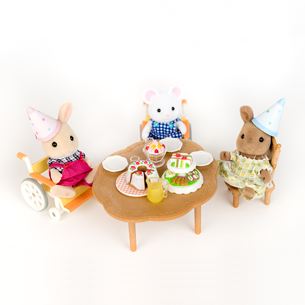 Image shows three Sylvanian figures having a tea party around a table. They are wearing party hats. Two figures are using wheelchairs.