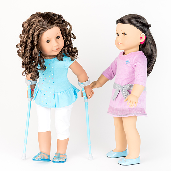 Two American Girl dolls. One with a hot pink hearing aid, the other with pale aqua crutches.