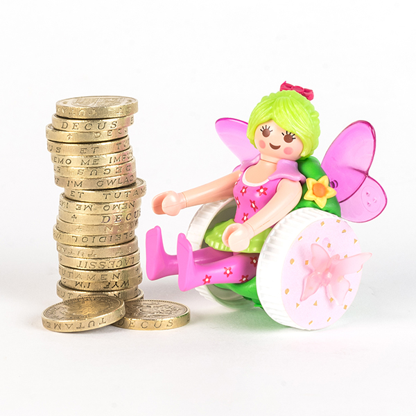 #ToyLikeMe makeover image of Playmobil fairy figure using a pink and green wheelchair made of modelling clay. The back of the chair has pink wings. She is sat next to a pile of one pound coins.