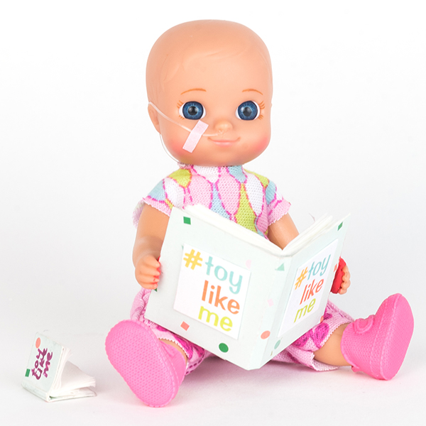 #ToyLikeMe makeover image showing baby girl toy with nasal feeding tube, reading a book.