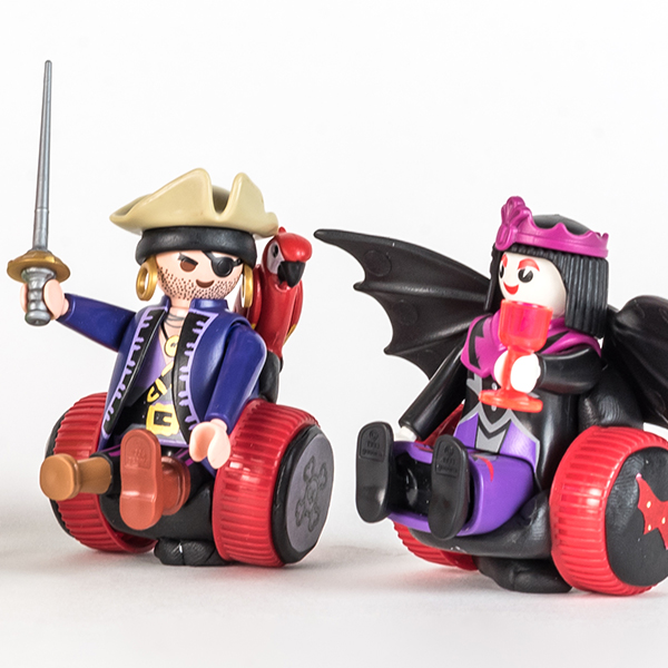 #ToyLikeMe makeover image of Playmobil Draculette figure using a red and black wheelchair made of modelling clay. The back of the chair has bat wings. She is sat next to a pirate figure using a red and black wheelchair.