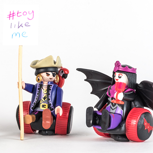#ToyLikeMe makeover image of Playmobil Draculette figure using a red and black wheelchair made of modelling clay. The back of the chair has bat wings. She is sat next to a pirate figure using a red and black wheelchair and carrying a sign reading 'toy like me'.