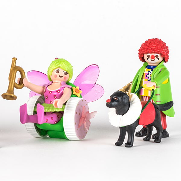 #ToyLikeMe makeover image of Playmobil clown figure with black guide dog. The dog is wearing a model clown's ruffle, gold pointy hat, red guide dog harness and has a red nose. There is also a fairy figure using a model wheelchair.